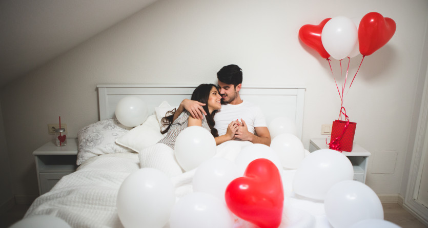 Couple allong&eacute; dans un lit, entour&eacute; de ballons rouges et blancs, dans une chambre romantique