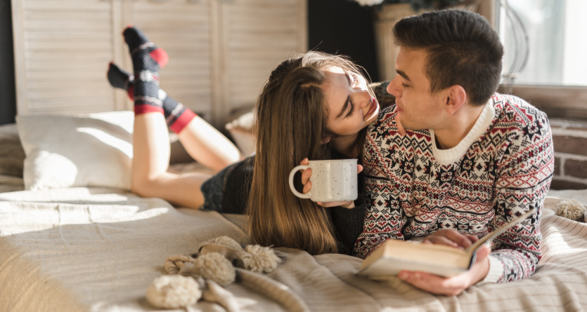 Couple en pull d'hiver partageant un moment cosy au lit avec un caf&eacute; et un livre
