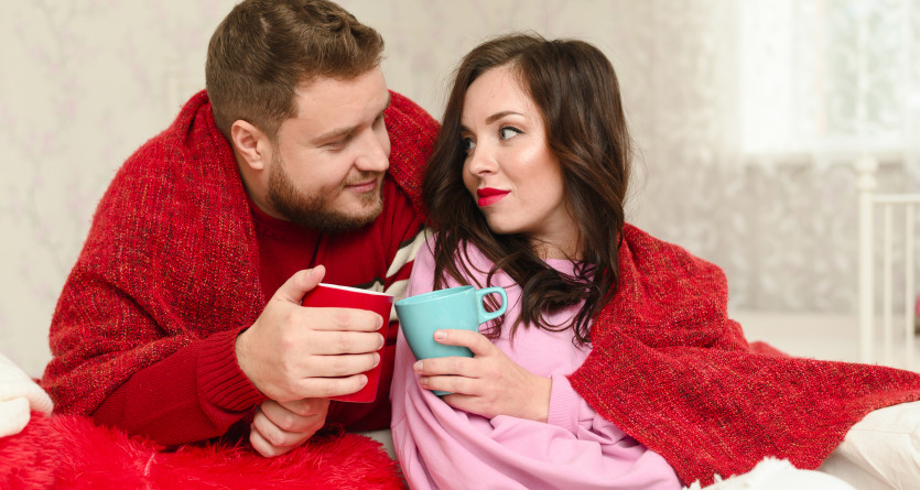 Un couple partage un moment chaleureux et complice dans une chambre romantique, envelopp&eacute;s dans des couvertures rouges.