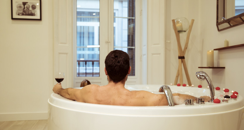 Homme savourant un verre de vin dans un jacuzzi parsem&eacute; de p&eacute;tales de roses, moment Love Room intime.