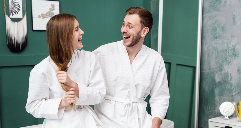 Un couple souriant en peignoirs blancs, se divertissant dans une salle de bain moderne avec des murs verts.