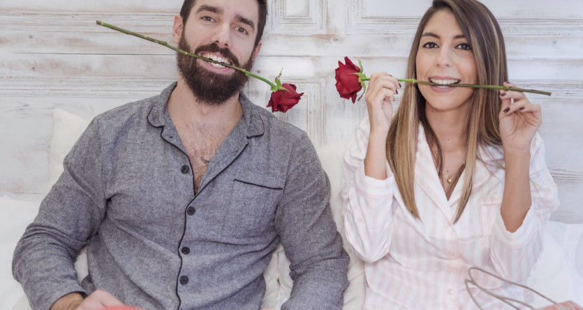 Couple joyeux tenant des roses rouges entre leurs dents dans une chambre lumineuse