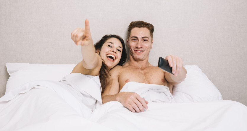 Couple joyeux regardant la t&eacute;l&eacute;vision au lit, partageant un moment de d&eacute;tente