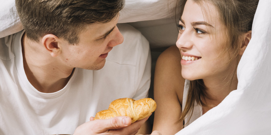 Couple &eacute;changeant un regard tendre sous les draps en d&eacute;gustant caf&eacute; et croissant