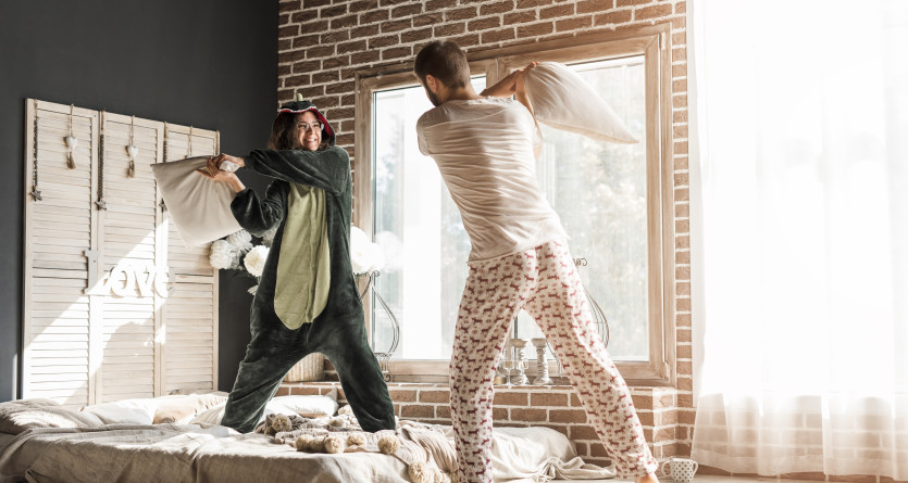 Couple s'amusant avec une bataille de polochons dans une chambre ludique et ensoleill&eacute;e