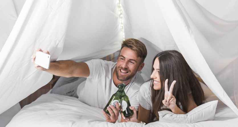 Couple allong&eacute; dans un lit sous un drap&eacute; blanc, prenant un selfie et partageant un moment complice