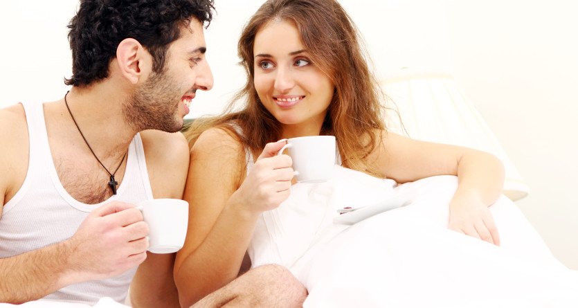 Couple souriant dans un lit, partageant un moment avec des tasses de caf&eacute;