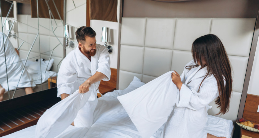 Couple en peignoirs blancs s'amusant dans une chambre d'h&ocirc;tel avec une bataille d'oreillers