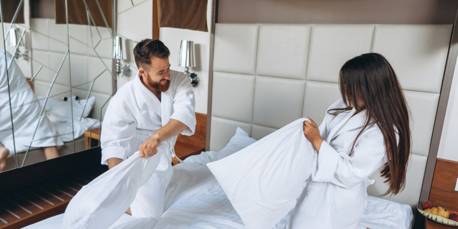 Couple en peignoirs blancs s'amusant dans une chambre d'h&ocirc;tel avec une bataille d'oreillers