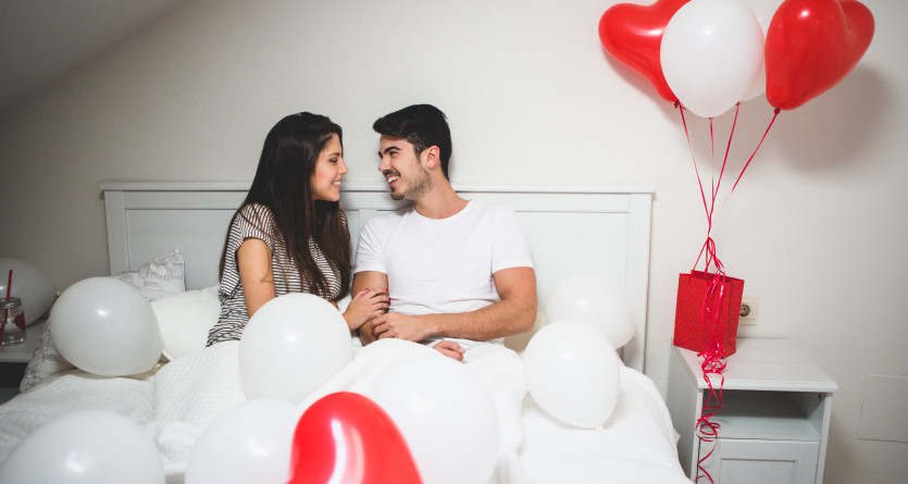 Couple souriant dans un lit entour&eacute; de ballons rouges et blancs en forme de c&oelig;ur